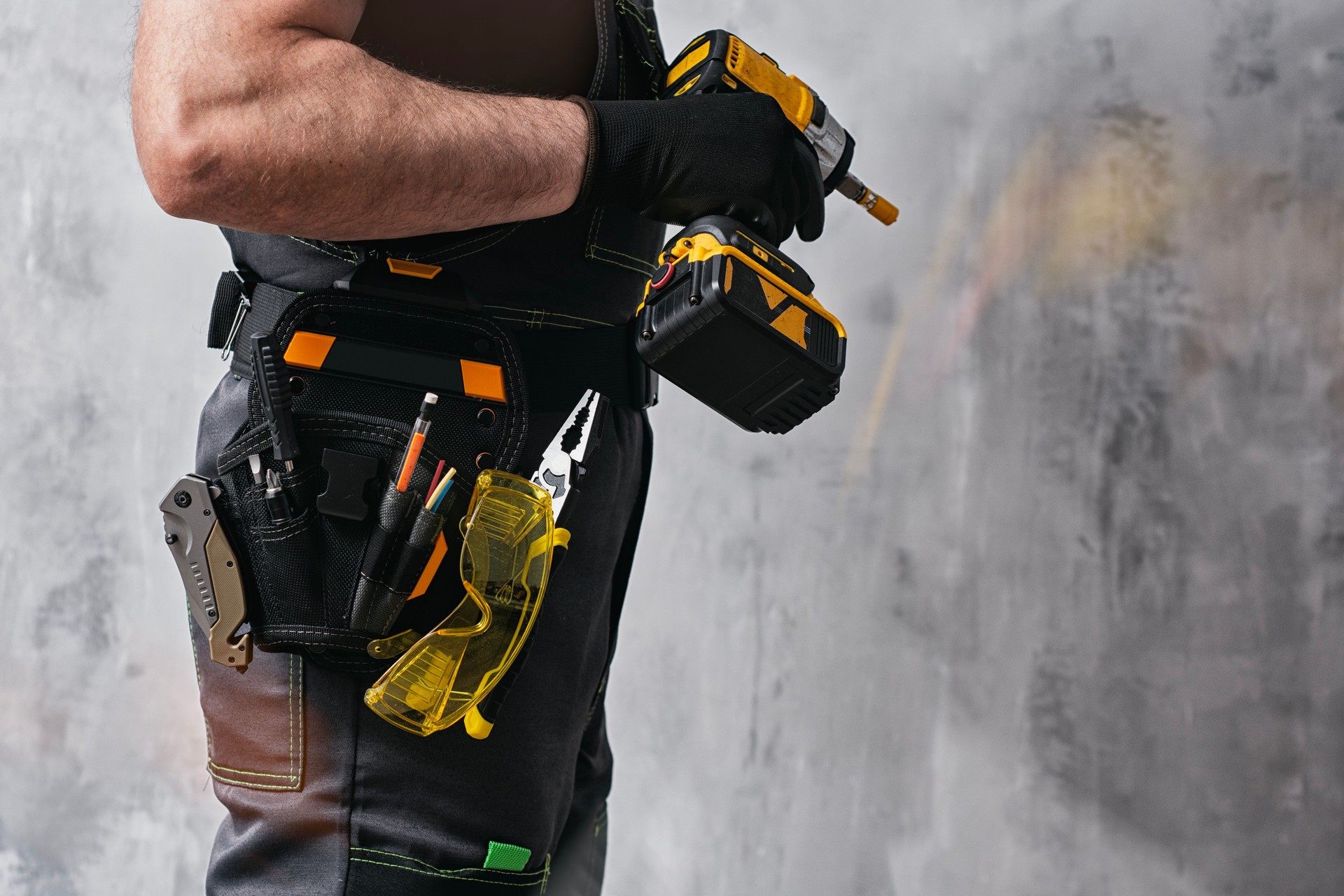 Uniformed construction worker holding a yellow colored cordless screwdriver with a set of tools on his belt. The concept of repair, maintenance and electrical installation.
